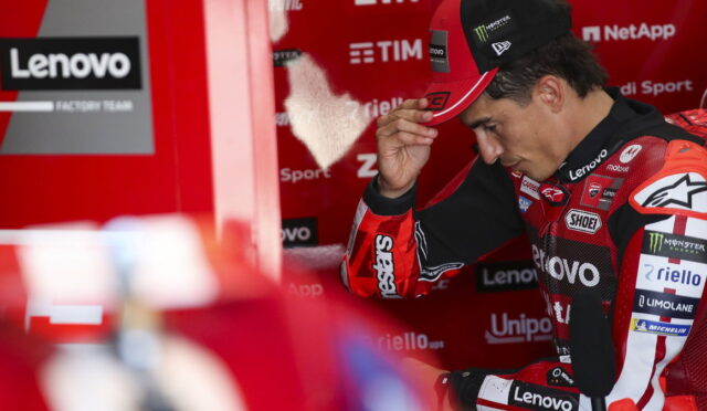 MotoGP rider in a red Lenovo racing suit adjusts his cap in the pit area.