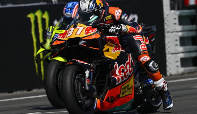 Motorcycle racer leaning into a turn on a KTM race bike with Red Bull livery, Monster energy backdrop in the background.