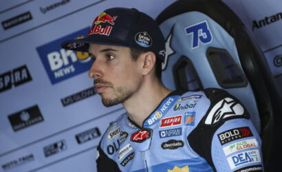 Motorcycle racer in a blue racing suit and cap with sponsor logos, seated in a race paddock with a backdrop of sponsor banners.