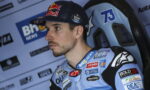 Male motorcycle racer wearing a Red Bull cap and blue racing suit with sponsor patches, seated in a paddock and looking off to the left.