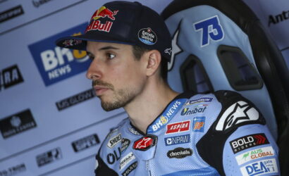 Male motorcycle racer wearing a Red Bull cap and blue racing suit with sponsor patches, seated in a paddock and looking off to the left.