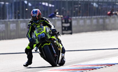 Motorcycle racer in a neon yellow-green suit leaning into a turn on a race track with a blue-red helmet.