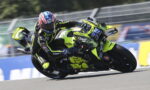 Two motorcycle racers lean deeply into a turn on a racetrack, neon-yellow bikes and matching gear dominating the scene.