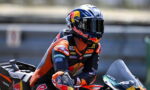 Moto racer in a Red Bull helmet leaning into a turn on an orange KTM race bike.