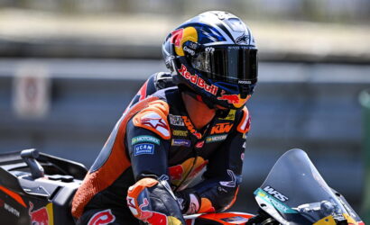 Moto racer in a Red Bull helmet leaning into a turn on an orange KTM race bike.