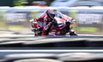 MotoGP rider in a red Ducati racing suit leans into a turn on a blurred race track edge.