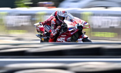 MotoGP rider in a red Ducati racing suit leans into a turn on a blurred race track edge.