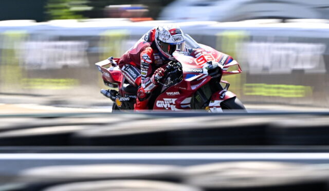 MotoGP rider in a red Ducati racing suit leans into a turn on a blurred race track edge.