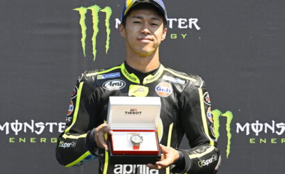 MotoGP rider in a black-yellow racing suit holds an open Tissot watch box, with a Monster Energy backdrop behind him.