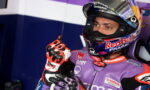MotoGP rider in a purple racing suit and Red Bull Pramac helmet in the pit, pointing a gloved finger upward.