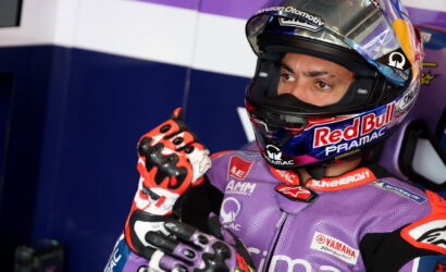 MotoGP rider in a purple racing suit and Red Bull Pramac helmet in the pit, pointing a gloved finger upward.