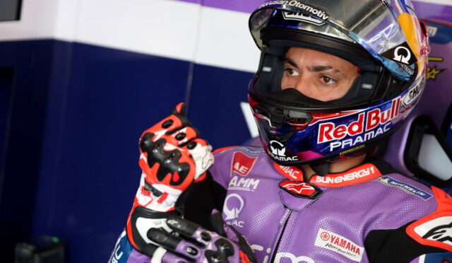 MotoGP rider in a purple racing suit and Red Bull Pramac helmet in the pit, pointing a gloved finger upward.