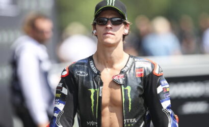 Moto racer in black Monster Energy jacket, cap, and sunglasses, standing outdoors with a blurred crowd in the background.