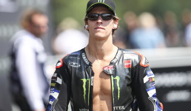 Moto racer in black Monster Energy jacket, cap, and sunglasses, standing outdoors with a blurred crowd in the background.