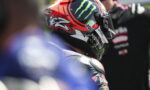 Racer wearing a red and black AGV helmet with Monster Energy logo, seen from the side in a pit area.