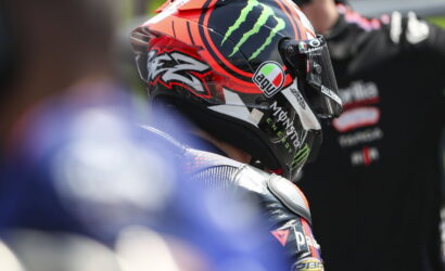 Racer wearing a red and black AGV helmet with Monster Energy logo, seen from the side in a pit area.