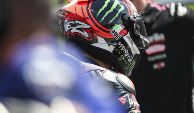 Racer wearing a red and black AGV helmet with Monster Energy logo, seen from the side in a pit area.