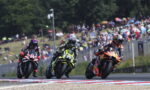 Three motorcycle racers lean into a tight turn on a crowded race track as spectators watch from the hillside and stands behind a fence.
