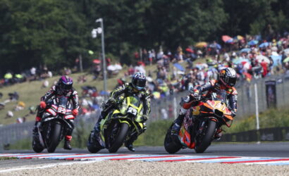 Three motorcycle racers lean into a tight turn on a crowded race track as spectators watch from the hillside and stands behind a fence.