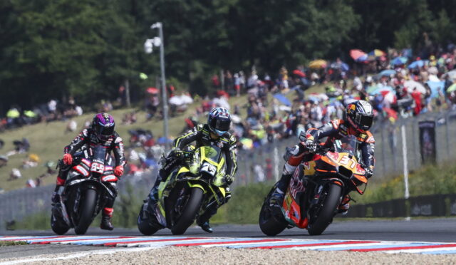 Three motorcycle racers lean into a tight turn on a crowded race track as spectators watch from the hillside and stands behind a fence.