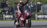 Racing rider on a red Ducati Lenovo bike raises a fist in celebration on a track, with Monster Energy banners in the background.