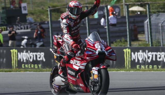 Racing rider on a red Ducati Lenovo bike raises a fist in celebration on a track, with Monster Energy banners in the background.