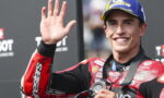 Motorcycle racer in red-and-black suit waves from the podium, wearing a blue Michelin cap and a medal around the neck.