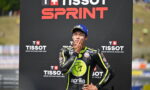 Race car/ motorcycle racer in a black and neon-yellow suit stands with hand on chin in front of a TISSOT SPRINT backdrop.