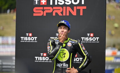 Race car/ motorcycle racer in a black and neon-yellow suit stands with hand on chin in front of a TISSOT SPRINT backdrop.