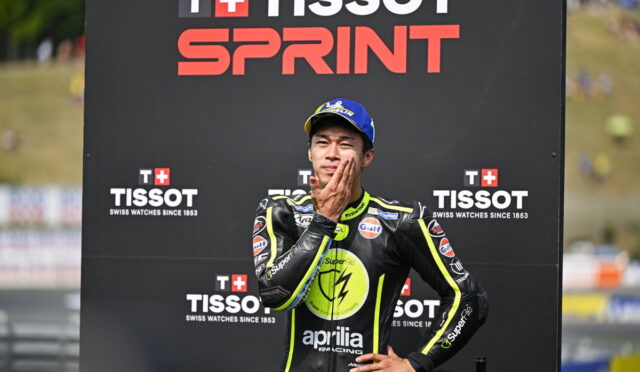 Race car/ motorcycle racer in a black and neon-yellow suit stands with hand on chin in front of a TISSOT SPRINT backdrop.