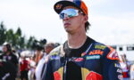 Motocross rider in blue/orange racing suit and white mirrored goggles, facing forward at the track with a crowd behind him.