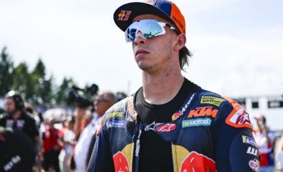 Motocross rider in blue/orange racing suit and white mirrored goggles, facing forward at the track with a crowd behind him.