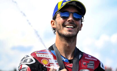 Male motorcycle racer in a red suit and cap, wearing sunglasses, smiling with a medal around his neck as champagne sprays in the background.