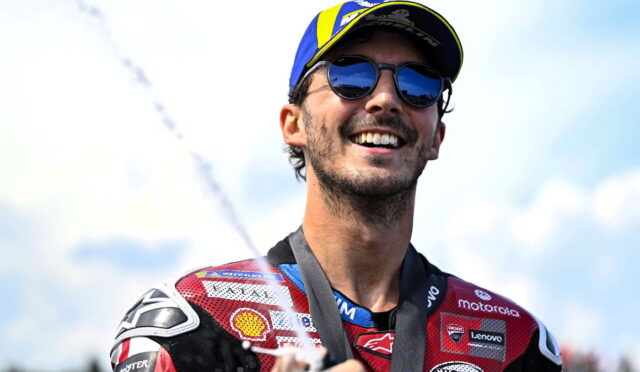 Male motorcycle racer in a red suit and cap, wearing sunglasses, smiling with a medal around his neck as champagne sprays in the background.