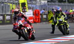 Racing motorcycles on a track; a red-and-white bike leads, rider leaning into a turn in red race gear, another yellow-and-black bike nearby.
