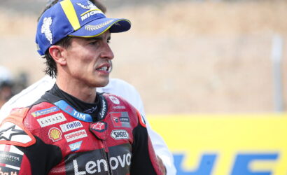 MotoGP rider wearing a red Lenovo racing suit and a blue/yellow Michelin cap on the track, looking focused with a clenched jaw