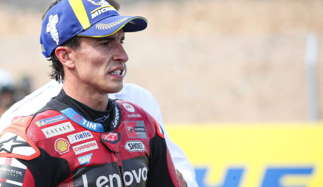 MotoGP rider wearing a red Lenovo racing suit and a blue/yellow Michelin cap on the track, looking focused with a clenched jaw