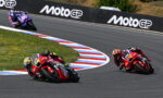 Three motorcycle racers lean into a sharp turn on a track, red and blue marked curbs visible, MotoGP barriers in the background.