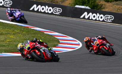 Three motorcycle racers lean into a sharp turn on a track, red and blue marked curbs visible, MotoGP barriers in the background.