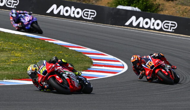 Three motorcycle racers lean into a sharp turn on a track, red and blue marked curbs visible, MotoGP barriers in the background.