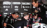MotoGP rider in racing suit and cap sits with three team members in a garage, reviewing notes with sponsor logos in the background.
