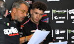 Two men in racing team polo shirts review papers at a backdrop of sponsor logos during a press event.