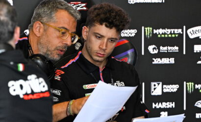 Two men in racing team polo shirts review papers at a backdrop of sponsor logos during a press event.