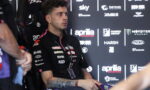 Young male motorbike racer in a black Aprilia Racing shirt sits in a chair during a press event, backdrop covered in sponsor logos shows his focused expression.
