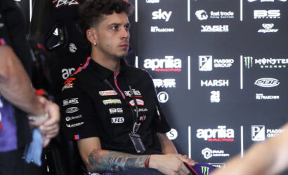 Young male motorbike racer in a black Aprilia Racing shirt sits in a chair during a press event, backdrop covered in sponsor logos shows his focused expression.