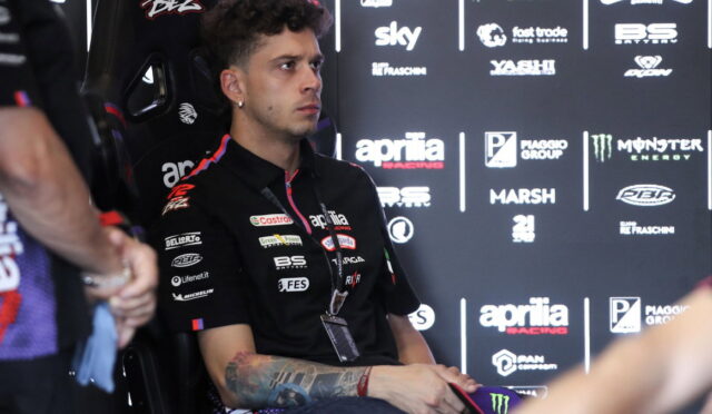 Young male motorbike racer in a black Aprilia Racing shirt sits in a chair during a press event, backdrop covered in sponsor logos shows his focused expression.