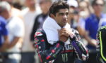 Male motorcycle racer with a white towel around his neck and a bandage on his nose, standing among spectators in the background.