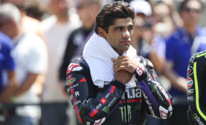 Male motorcycle racer with a white towel around his neck and a bandage on his nose, standing among spectators in the background.