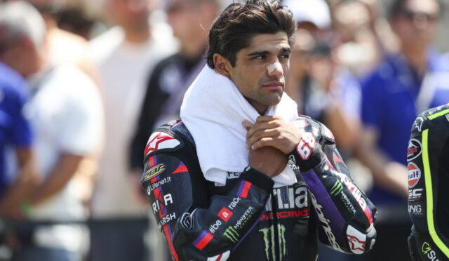 Male motorcycle racer with a white towel around his neck and a bandage on his nose, standing among spectators in the background.