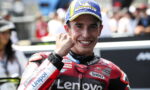 MotoGP rider in a red Lenovo racing suit, smiling with a raised fist, wearing a blue Michelin cap; cheering crowd in the background.